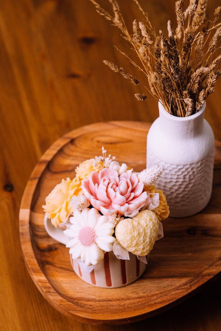 herzton Blumentasse mit gelben und rosa Wachsblüten auf rundem Holztablett, Trockenblumen-Vase im Hintergrund