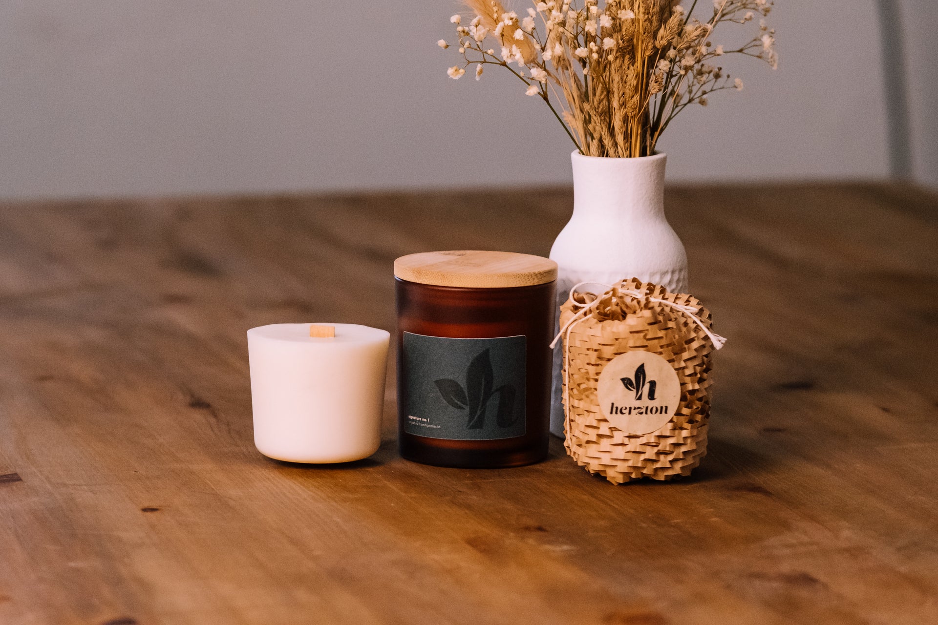 herzton Sojawachskerze im Amberfarbglas mit Holzdeckel, kleine weiße Kerze und Bast-Geschenkbeutel auf Holz