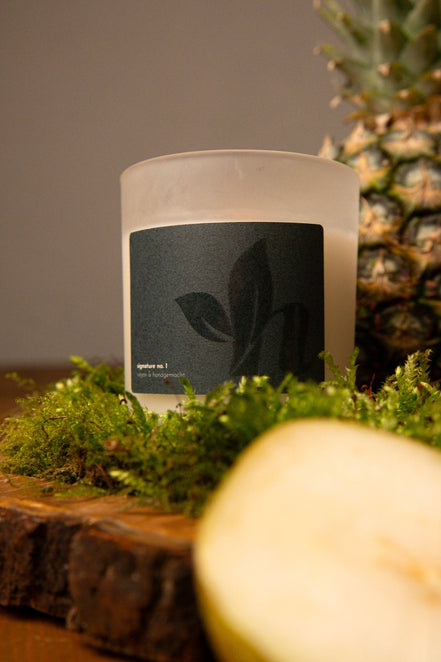 herzton Signature Duftkerze im satinierten Milchglas mit dunklem Etikett, auf Holz mit Ananas und Moos