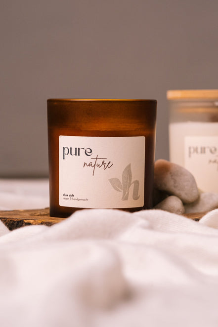 herzton Pure Nature Sojawachskerze im Bernsteinglas mit weißem Etikett "pure nature – ohne duft", auf Holz mit Steinen