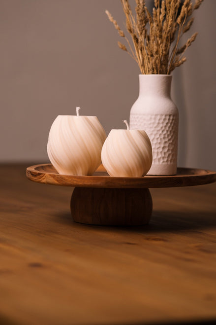 herzton Wave Duo, zwei cremeweiße Kugelkerzen mit Wellenmuster auf Holzsockel, Trockenblumen-Vase dahinter